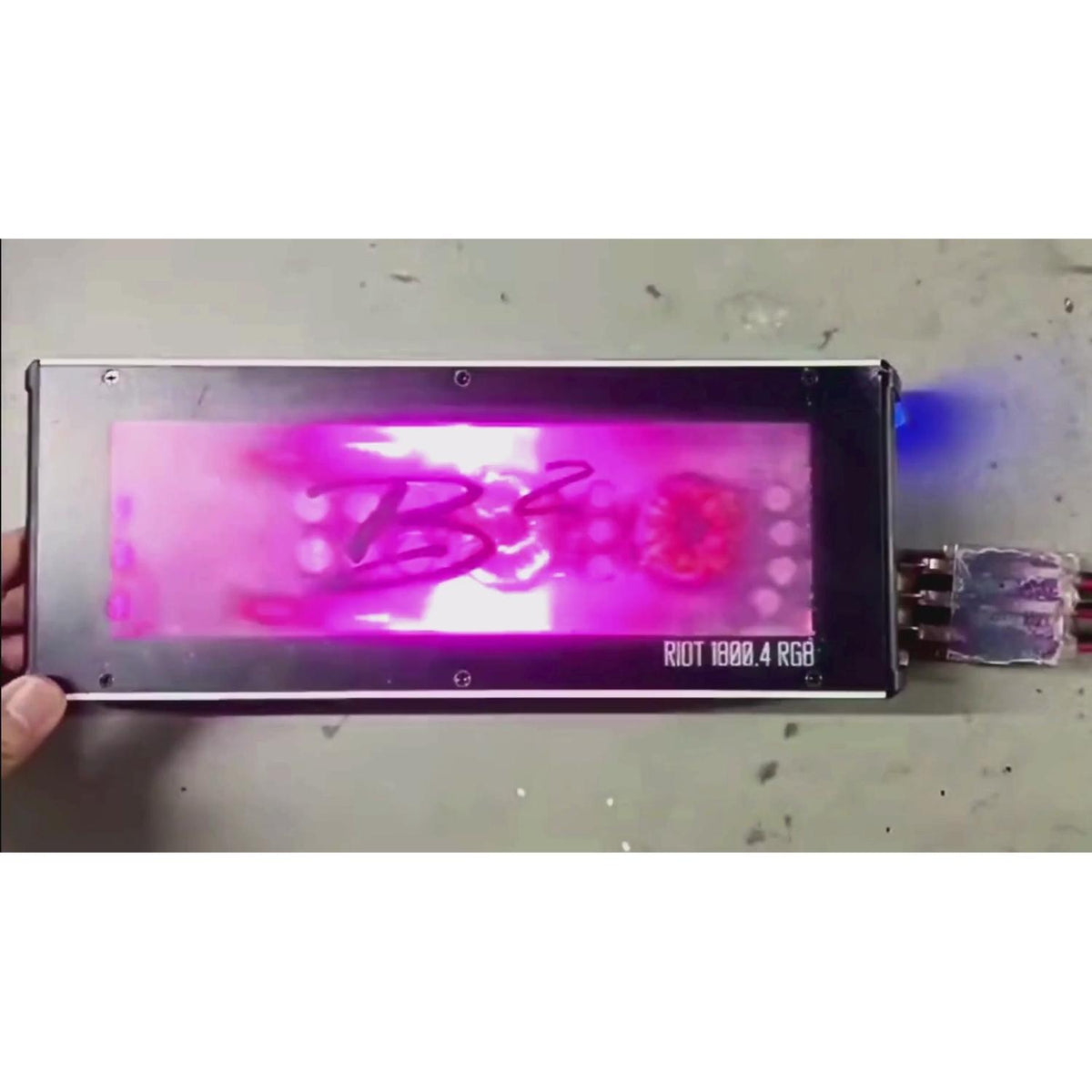 Un amplificateur rectangulaire à 4 canaux B2 Audio Riot 1800.4RGB, doté dun écran lumineux rose-violet, affiche « B340 » et « RIOT 1800.4 RGB » en bas à droite ; une main apparaît à gauche.