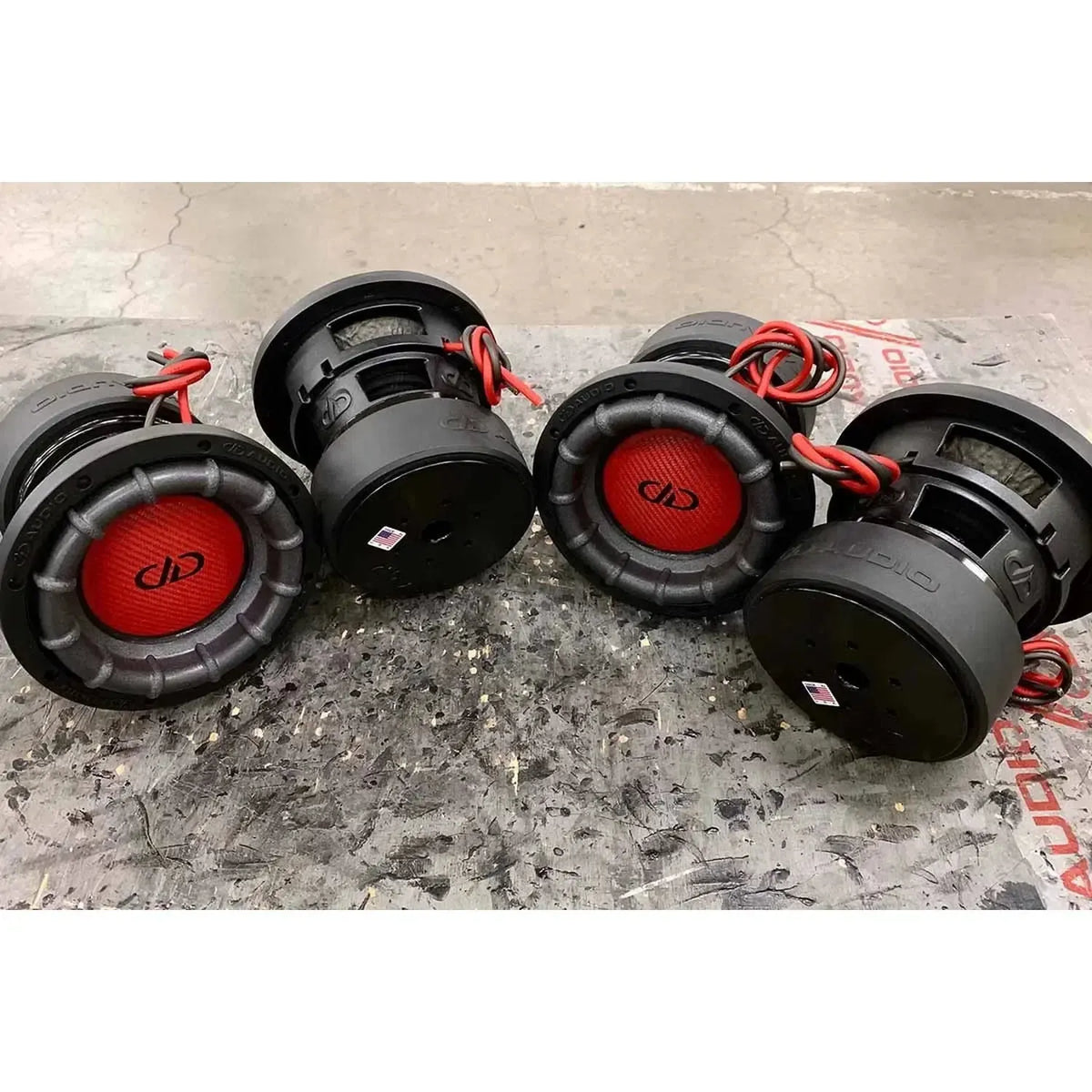 Vier grote DD Audio Power Tuned 1506 ESP subwoofers, 600W RMS elk met rode draden, staan op een stoffige ondergrond, in verschillende richtingen.