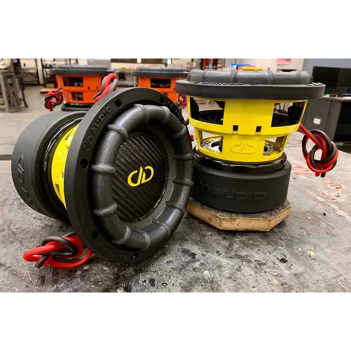 Twee DD Audio Power Tuned 1506 ESP subwoofers, 5, met zwarte koolstofvezel conussen, gele frames, rode draden, 600W RMS  dubbele spoelen van 2 ohm, staan op een werktafel.