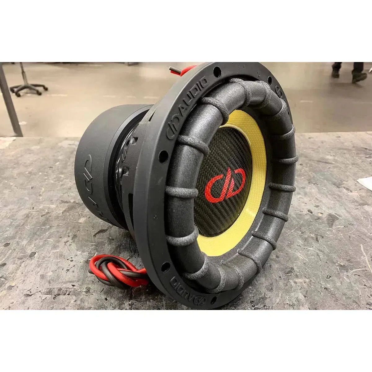 Un subwoofer DD Audio Power Tuned 2508F ESP nero e giallo con logo rosso è appoggiato su un banco da lavoro grigio con fili rossi e neri attaccati.