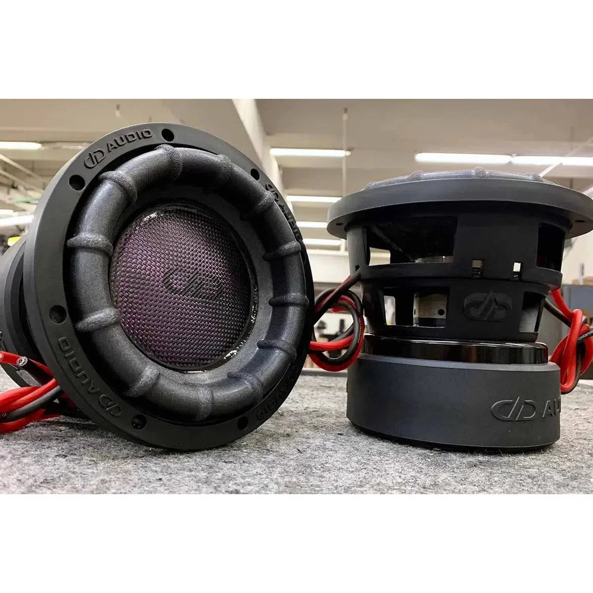 Due subwoofer DD Audio Power Tuned 2508F ESP con cablaggio rosso sono collocati su una superficie grigia: uno in posizione verticale, laltro su un fianco.