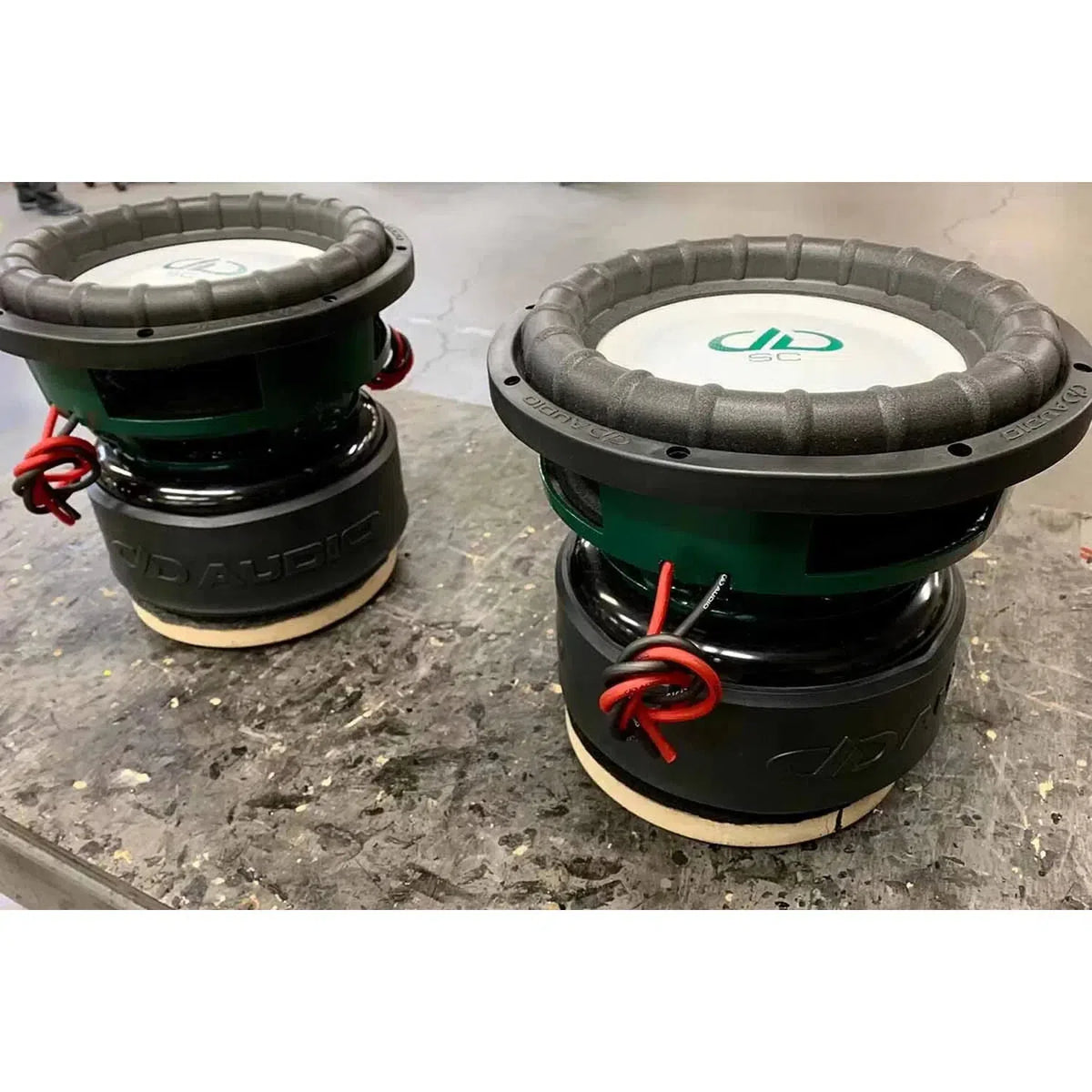 Two large DD Audio Power Tuned 9512k ESP subwoofers with carbon fiber blend cones and red wires sit on a gray, slightly worn workbench.