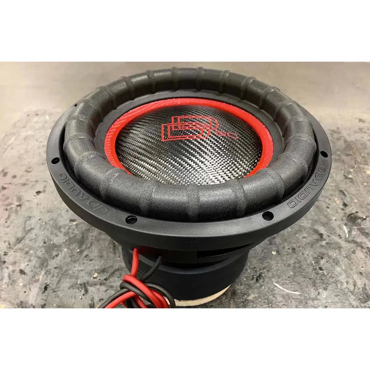 Un subwoofer DD Audio Power Tuned 9912b ESP da 12 con cono in fibra di carbonio, design nero e rosso e logo centrale si trova su una superficie grigia con fili rossi visibili.