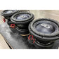 Drie grote DD Audio Power Tuned 9915b ESP subwoofers met koolstofvezel mengkegels en rode draden staan op een rij op een zwart werkoppervlak binnenshuis.