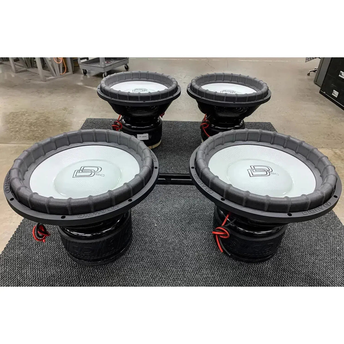 Vier DD Audio Power Tuned 9915b ESP 15 subwoofers met koolstofvezel mengkegels en zwarte frames worden op een grijs tapijt in een werkplaats geplaatst.