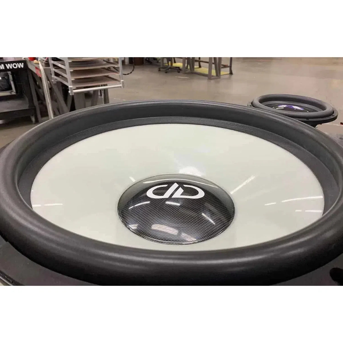 Close-up van de DD Audio Power Tuned 9921b 21-inch subwoofer met zwart-witte conus, middenstuk met koolstofvezelpatroon en ferrietmagneet in een industriële werkplaats.