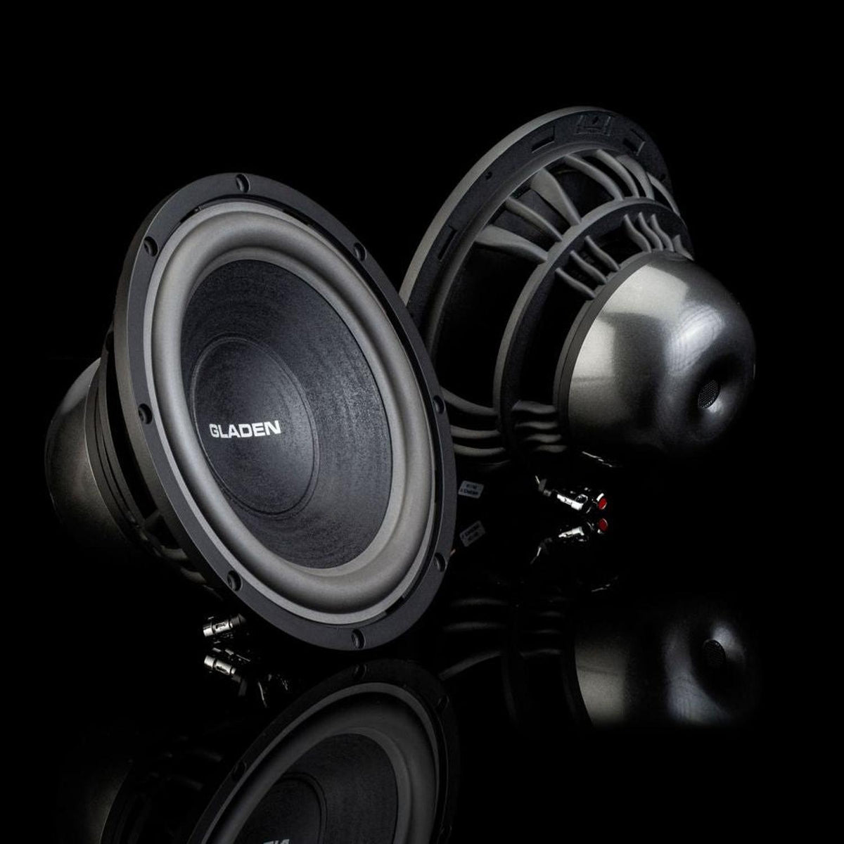 Twee zwarte Gladen Aerospace Aero 10-subwoofers staan op een reflecterend oppervlak, één naar voren gericht en één naar achteren, waarmee ze hun hoogwaardige Auto Hifi-stijl laten zien.