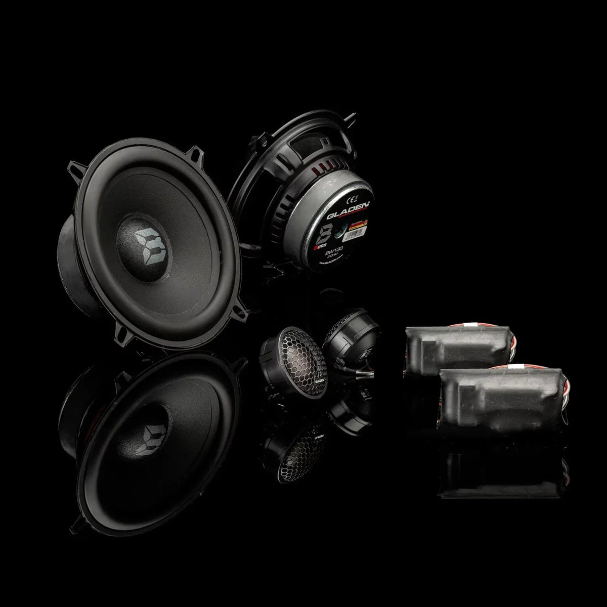 The Gladen Beta 130.2 features two large speakers, a tweeter, and crossovers, all showcased on a glossy black surface with striking reflections.