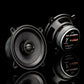 Two Gladen Gamma 130-X speakers on a glossy black surface, one front-facing, one showing the back label, both with sleek reflections.