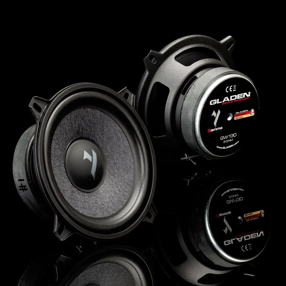 Deux enceintes Gladen Gamma 130.2 noires sont présentées sur une surface réfléchissante : lune montre son cône et lautre son étiquette arrière, mettant ainsi en valeur leur qualité sonore.