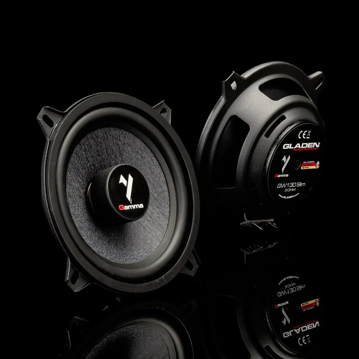 Two round black Gladen Gamma 130.2 Slim speakers are shown on a glossy surface—one faces forward, the other reveals its back.