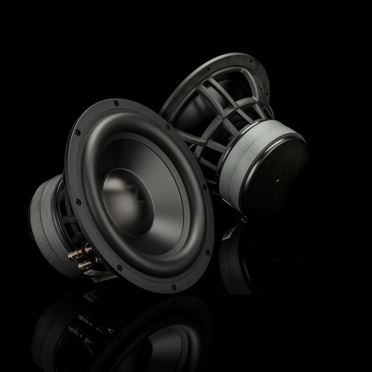 Deux caissons de basses Gladen Zeta 10 diffusent des basses puissantes et sont posés sur une surface noire brillante, devant un arrière-plan sombre et minimaliste.